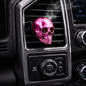 May include: A shiny pink skull-shaped car air freshener is positioned in a car's air vent. The skull has detailed features. The car's dashboard is dark grey with control knobs and buttons.