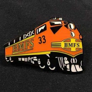 May include: An orange and black train-shaped pin featuring the text "BMFS" and the number "33". The pin has yellow and white detailing and is set against a dark background.