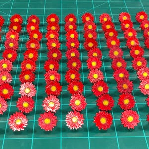 Op de afbeelding: Een raster van kleine, rode papieren bloemen met gele centra. De bloemen zijn in nette rijen gerangschikt op een groene snijmat. De bloemen zijn gelijkmatig verdeeld en lijken handgemaakt.