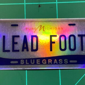 May include: A rectangular sticker resembling a license plate, featuring the text "LEAD FOOT" in bold, dark blue letters. The top of the sticker reads "PURE MICHIGAN" in a stylized font. The bottom reads "BLUEGRASS". The background has a rainbow holographic effect.