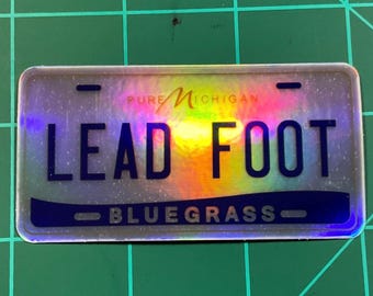 May include: A rectangular sticker resembling a license plate, featuring the text "LEAD FOOT" in bold, dark blue letters. The top of the sticker reads "PURE MICHIGAN" in a stylized font. The bottom reads "BLUEGRASS". The background has a rainbow holographic effect.