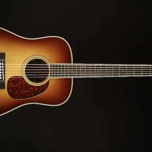 May include: Acoustic guitar with a sunburst finish, featuring a brown and tan gradient on the body. The guitar has a round sound hole, a tortoiseshell pickguard, and a dark fretboard. The headstock displays decorative inlays and tuning pegs.
