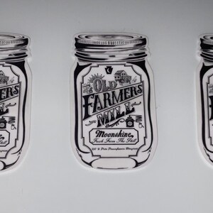 Op de afbeelding: Drie zwart-witte stickers in de vorm van weckpotten. Elke sticker toont de tekst "Old Farmers Mill Moonshine" in een vintage stijl, met illustraties van een huis en een distilleerketel.