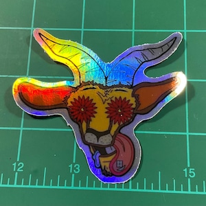 May include: Holographic sticker featuring a cartoon goat with red flower eyes and a lolling tongue. The goat has rainbow-colored horns and ears, with a yellow and brown face. The sticker is on a green surface with measurement markings.