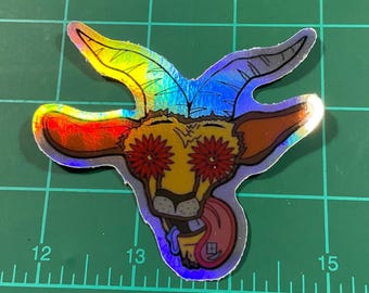 May include: Holographic sticker featuring a cartoon goat with red flower eyes and a lolling tongue. The goat has rainbow-colored horns and ears, with a yellow and brown face. The sticker is on a green surface with measurement markings.