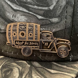 May include: A bronze-coloured pin depicting a vintage truck with a wooden barrel. The pin features the text "Must Be Seven" and the number "93". The pin is set against a camouflage patterned background.