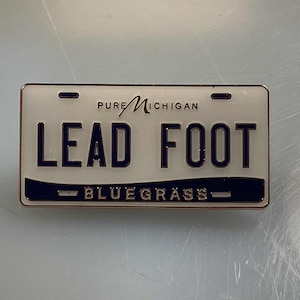May include: A rectangular metal license plate-shaped pin with a white background and black text that reads "LEAD FOOT" and "BLUEGRASS". The top of the pin reads "PURE MICHIGAN".