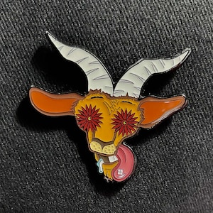Op de afbeelding: Een kleurrijke emaille pin met een geitenkop met witte hoorns, oranje oren en rode bloemogen. De geit steekt zijn tong uit en heeft een roze en witte tong. De pin staat tegen een donkere achtergrond.
