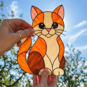 May include: A stained glass suncatcher shaped like a sitting cat with orange, cream, and brown glass segments. The cat features large black eyes, a pink nose, and thin black wire whiskers, held up against a bright blue sky with sunlight shining through.
