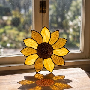 May include: A stained glass sunflower suncatcher featuring textured yellow petals and a dark brown centre, constructed with black metal lead lines. The piece is circular in shape and displays light transmission through the glass segments.