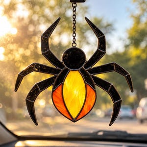 May include: A stained glass spider ornament hanging from a metal chain. The spider features eight black glass legs and a black head, with a central abdomen made of translucent yellow and orange glass panels held together by dark metal solder lines.