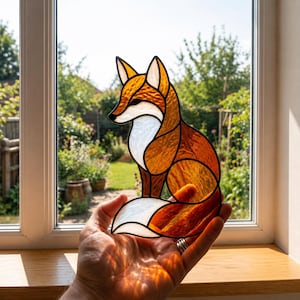 Op de afbeelding: Een glas-in-lood zonnevanger in de vorm van een vos, gemaakt van getextureerd amberkleurig en oranje glas voor het lichaam en de oren, met ondoorzichtig wit glas voor de borst en de staartpunt. De delen zijn verbonden door donkere metalen soldeerlijnen.