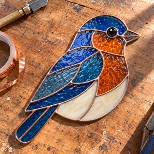 Puede incluir: Un atrapasoles de vidrieras con forma de pájaro que presenta vidrio azul texturizado en la cabeza, las alas y la cola, un pecho naranja vibrante y un vientre color crema. Las piezas de vidrio están unidas con finas líneas de soldadura de cobre.