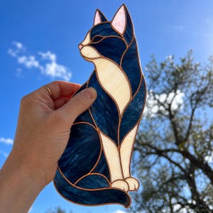 Peut inclure: Un attrape-soleil en vitrail représentant un chat, composé de pièces de verre bleu foncé et crème assemblées par des lignes de soudure en métal cuivré. Le chat est représenté de profil, assis, avec des oreilles pointues et un design segmenté.