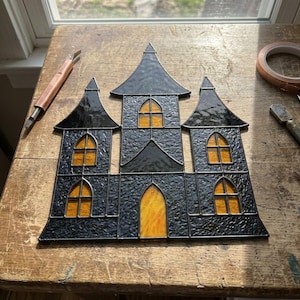 May include: A stained glass haunted house panel featuring textured black glass walls and roofs with glowing amber yellow glass windows and a central arched doorway, constructed with dark metal solder lines in a classic gothic architectural style.