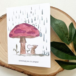 May include: A watercolor illustration on a white card features a mouse sheltering under a mushroom from the rain. The mushroom has a maroon cap and a brown stem. The text "covering you in prayer" is at the bottom of the card.