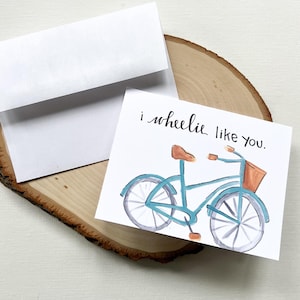 May include: A greeting card featuring a watercolour illustration of a blue bicycle with a brown seat and basket. The card reads "i wheelie like you." A white envelope is included.