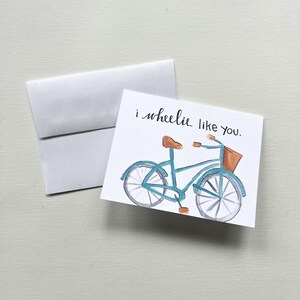 May include: A watercolour greeting card featuring a blue bicycle illustration and the phrase "i wheelie like you." The card has a brown seat and basket, and is accompanied by a white envelope.
