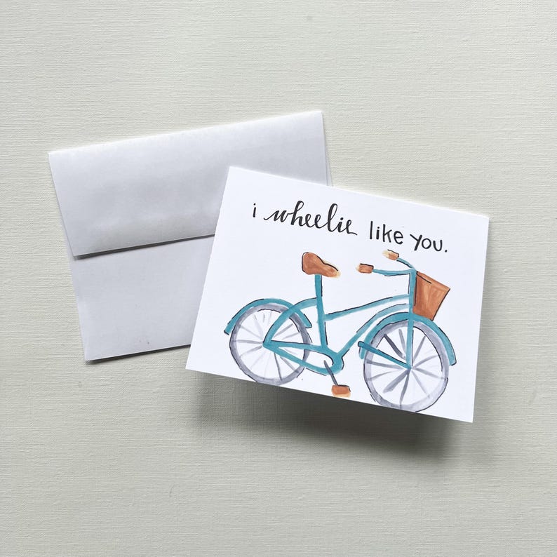 May include: A watercolour greeting card featuring a blue bicycle illustration and the phrase "i wheelie like you." The card has a brown seat and basket, and is accompanied by a white envelope.