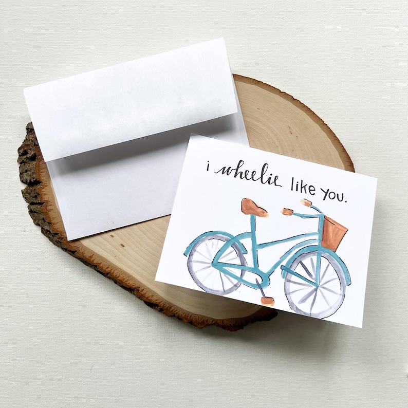May include: A greeting card featuring a watercolour illustration of a blue bicycle with a brown seat and basket. The card reads "i wheelie like you." A white envelope is included.