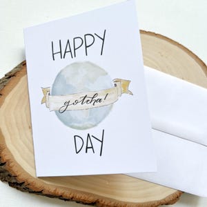 May include: A white greeting card with the words "HAPPY DAY" in black script. A light blue globe with a banner that reads "gotcha!" is in the center. An envelope is next to the card.