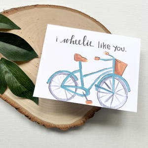 May include: A greeting card featuring a watercolour illustration of a blue bicycle with a brown seat and basket. The card has the text "i wheelie like you" in black script. The card is on a wooden slice with green leaves.