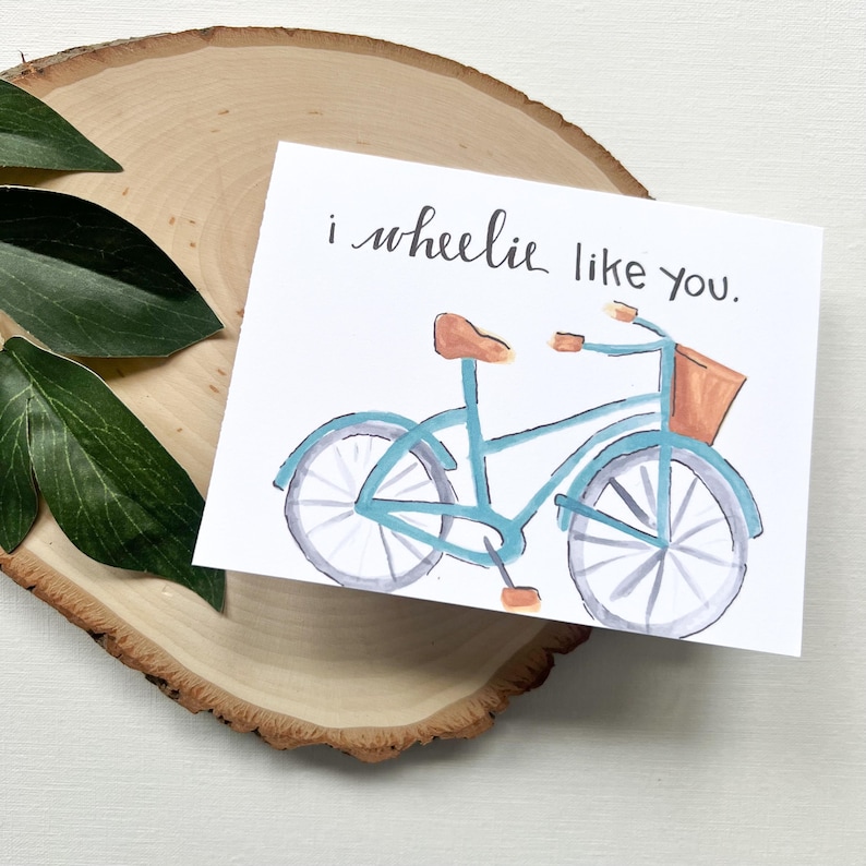 May include: A greeting card featuring a watercolour illustration of a blue bicycle with a brown seat and basket. The card has the text "i wheelie like you" in black script. The card is on a wooden slice with green leaves.