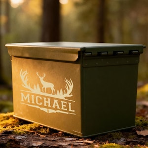 May include: An olive green metal storage box with a hinged lid. The box features a white graphic of a deer and the name "MICHAEL" in a decorative font. The box is sitting on a bed of moss and leaves in a forest setting.