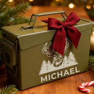 May include: An olive green metal box with a handle, decorated with a white deer and tree design, and the name "MICHAEL". A burgundy bow adorns the top. The box is on a wooden surface, surrounded by Christmas decorations.