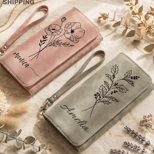 May include: Two suede-like wallets, one dusty rose and one gray, each with a wrist strap. The dusty rose wallet features a floral design and the name "Monica". The gray wallet has a botanical design and the name "Amelia".