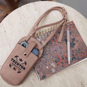 May include: A dusty rose-colored phone crossbody bag with a shoulder strap, holding a phone and a small device. The bag has the text "TRENTON - POWERLIFTING" printed on it. A floral notebook and a pen are next to the bag.