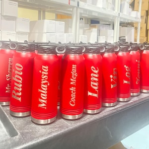 A row of bright red insulated water bottles with engraved personalized names stand on a dark surface in front of storage shelves.