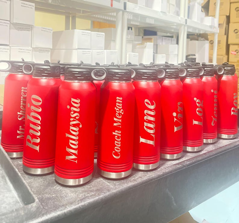A row of bright red insulated water bottles with engraved personalized names stand on a dark surface in front of storage shelves.
