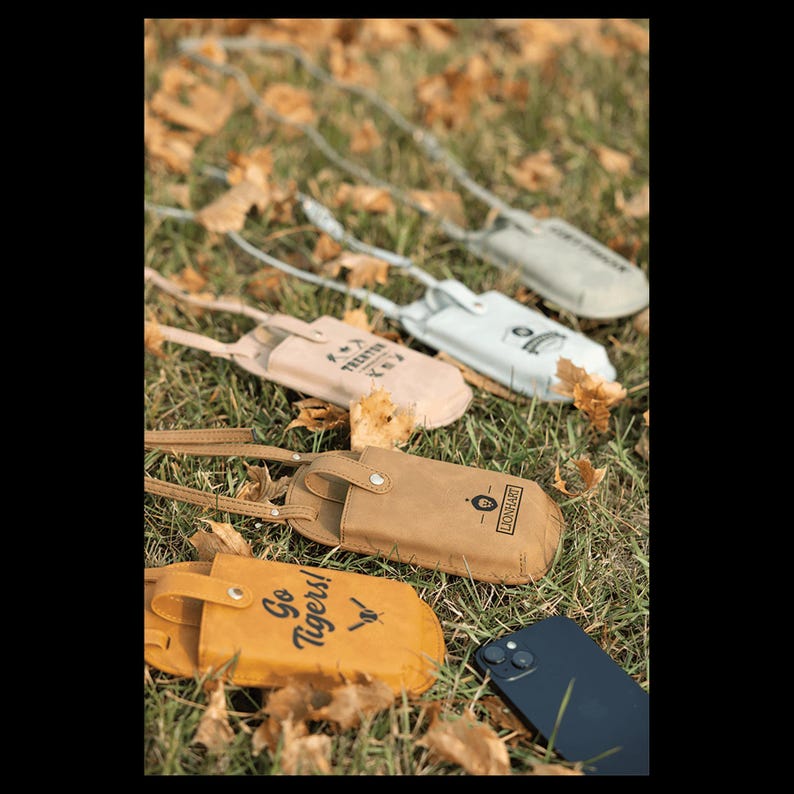 May include: A collection of leather phone cases in various colors, including tan, yellow, and white, with attached straps. Some cases feature text like "Go Tigers!" and "TECHTOX". The cases are displayed on grass with fallen leaves.