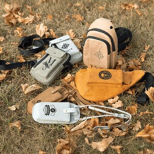 May include: An assortment of leather bags and accessories in various shades of tan, beige, and white, arranged on a bed of dry grass and fallen leaves. The collection includes a backpack, fanny pack, and several small pouches, some with printed designs.