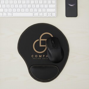 May include: Black ergonomic mouse pad with a wrist rest, a black wireless mouse, and a white keyboard. The mouse pad has a gold logo and the text "COMPA AGLINE GOES". A black smartphone is also visible.