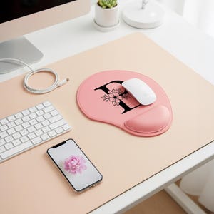 May include: A pink ergonomic mouse pad with a black letter "F" and floral design. A white mouse rests on the pad, next to a matching pink wrist rest. A white keyboard and a smartphone with a flower image are also on the desk.