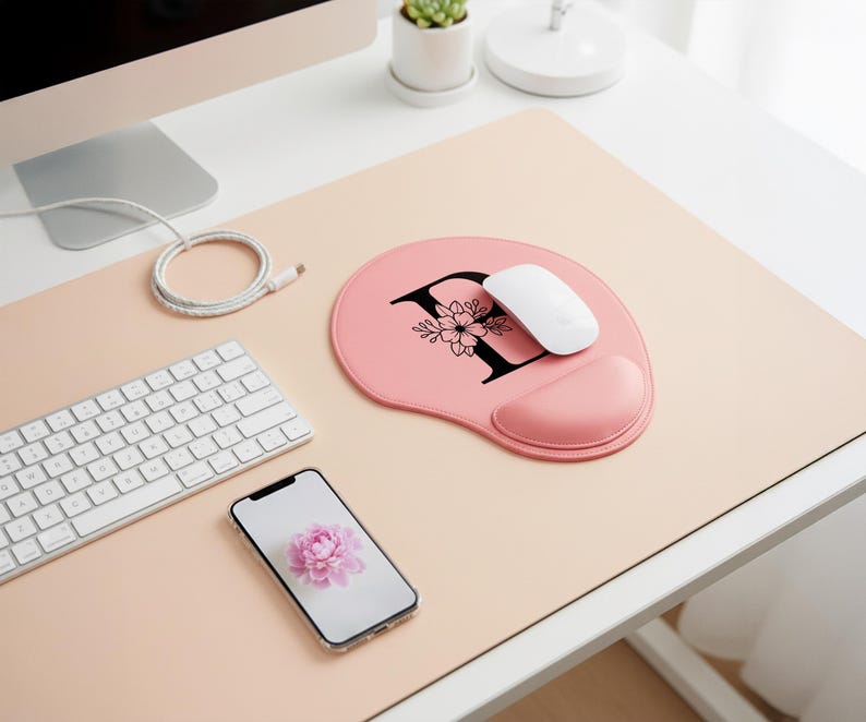 May include: A pink ergonomic mouse pad with a black letter "F" and floral design. A white mouse rests on the pad, next to a matching pink wrist rest. A white keyboard and a smartphone with a flower image are also on the desk.
