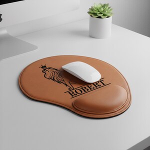 May include: A brown leather mouse pad with a black lion logo and the name "ROBERT". The mouse pad has a wrist rest and a white wireless mouse on top. A small succulent plant in a white pot is in the background.