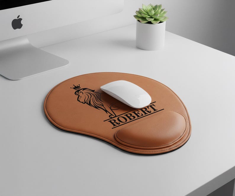 May include: A brown leather mouse pad with a black lion logo and the name "ROBERT". The mouse pad has a wrist rest and a white wireless mouse on top. A small succulent plant in a white pot is in the background.
