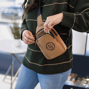 May include: A tan crossbody bag with a black zipper and a black floral design with the letter "K". The bag has a small front pocket and a long strap. The person is wearing a green sweater and blue jeans.
