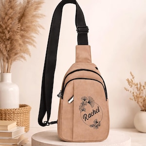 May include: A tan sling bag with a black strap and zipper details. The bag features a floral design with the name "Rachel" in a circular frame. The bag is displayed on a white pedestal.