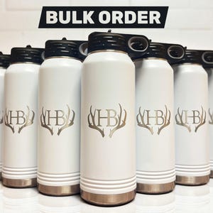 A group of matte white water bottles with black lids standing in rows, each engraved with the monogram letters HB surrounded by antlers.