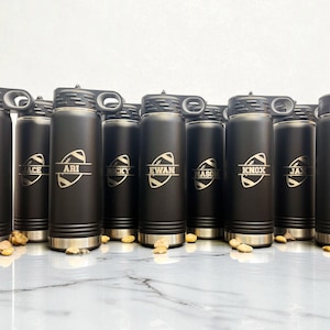 Puede incluir: Una colecci&oacute;n de botellas de agua aisladas negras con detalles plateados. Cada botella presenta un gr&aacute;fico de f&uacute;tbol americano y un nombre en blanco. Las botellas tienen una tapa negra con asa.