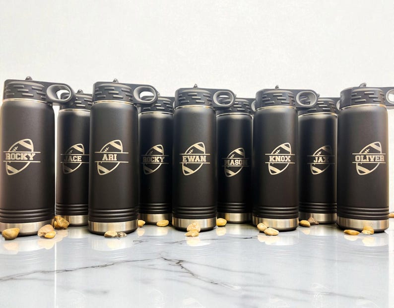 Puede incluir: Una colecci&oacute;n de botellas de agua aisladas negras con detalles plateados. Cada botella presenta un gr&aacute;fico de f&uacute;tbol americano y un nombre en blanco. Las botellas tienen una tapa negra con asa.