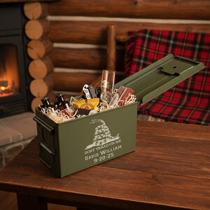 May include: An olive green metal ammo box, open to reveal a collection of gifts. The box features the text "DON'T TREAD ON ME" and a snake graphic. Inside are small bottles, snacks, and a pocket knife. The box sits on a wooden table.