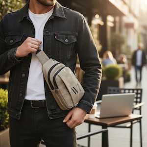 May include: A beige sling bag with black accents and the name "WILLIAM" printed vertically. The bag has a zippered front pocket and a black strap. The person is wearing a black denim jacket and black pants.