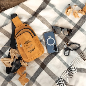 May include: Mustard yellow sling bag with a black strap, a front pocket, and the name "Sarah" printed on it. The bag is next to a phone in a blue case, sunglasses, and a keychain with a black beaded bracelet. The items are on a plaid blanket with autumn leaves.
