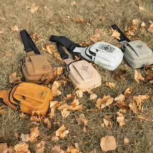 May include: A collection of crossbody bags in various colors, including tan, mustard yellow, and gray, are displayed on a bed of dry grass and fallen leaves. Each bag features a different design, such as a mountain, a compass, or a sun with the text "LUNA STAR".