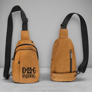 May include: Two tan sling bags with black straps. One bag has a front pocket and the text "DOG mom" with a paw print design. The other bag has a vertical zipper. Both bags have a black adjustable strap.